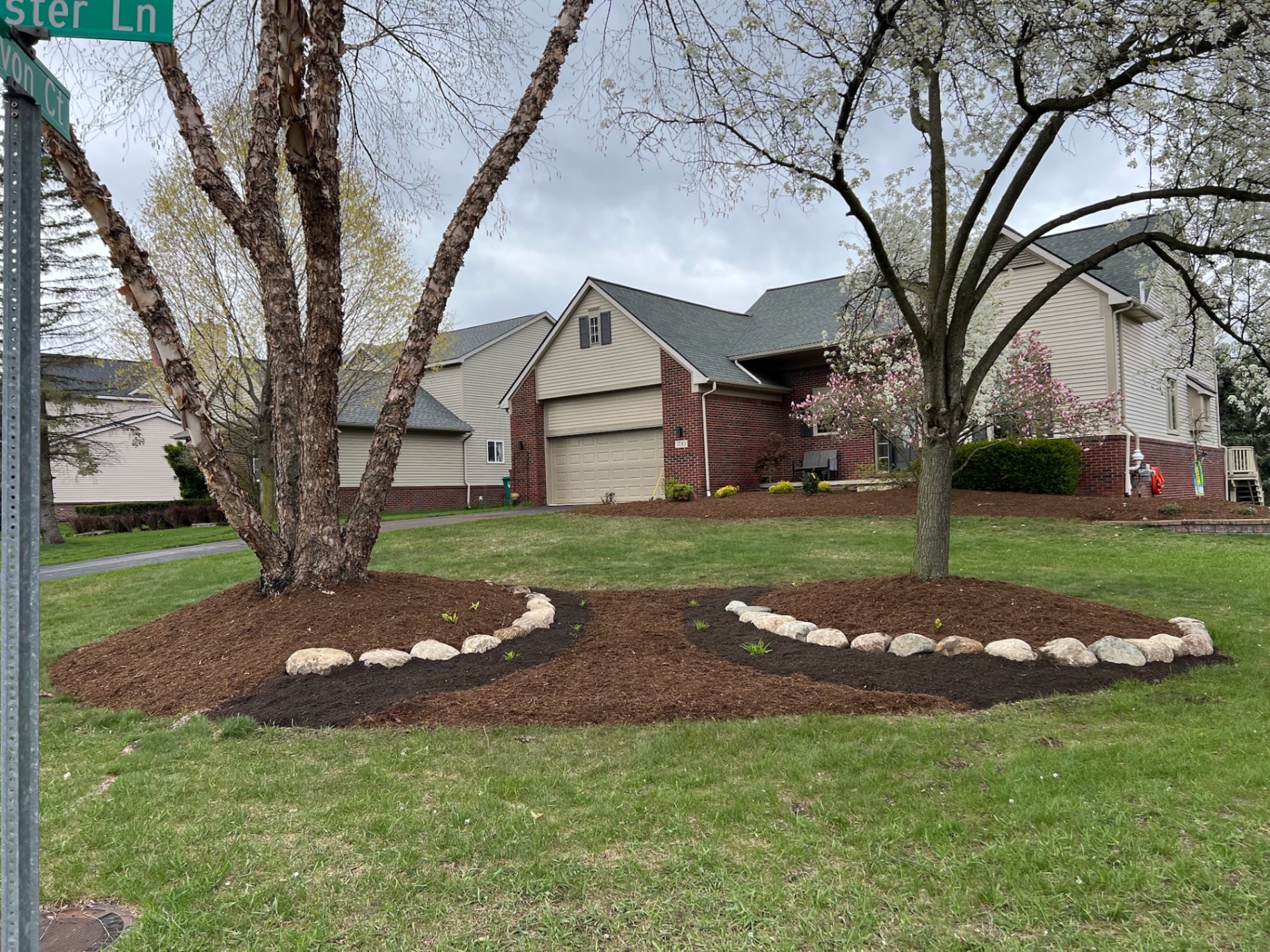 Curved planting beds connected with natural stone borders beneath mature front-yard trees.