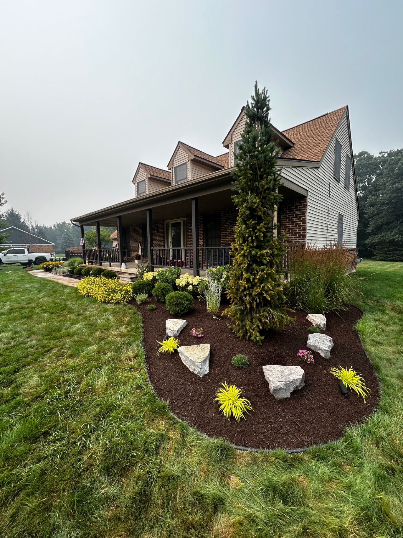 Dramatic front-yard island bed featuring an evergreen focal tree, accent boulders, and fresh mulch.